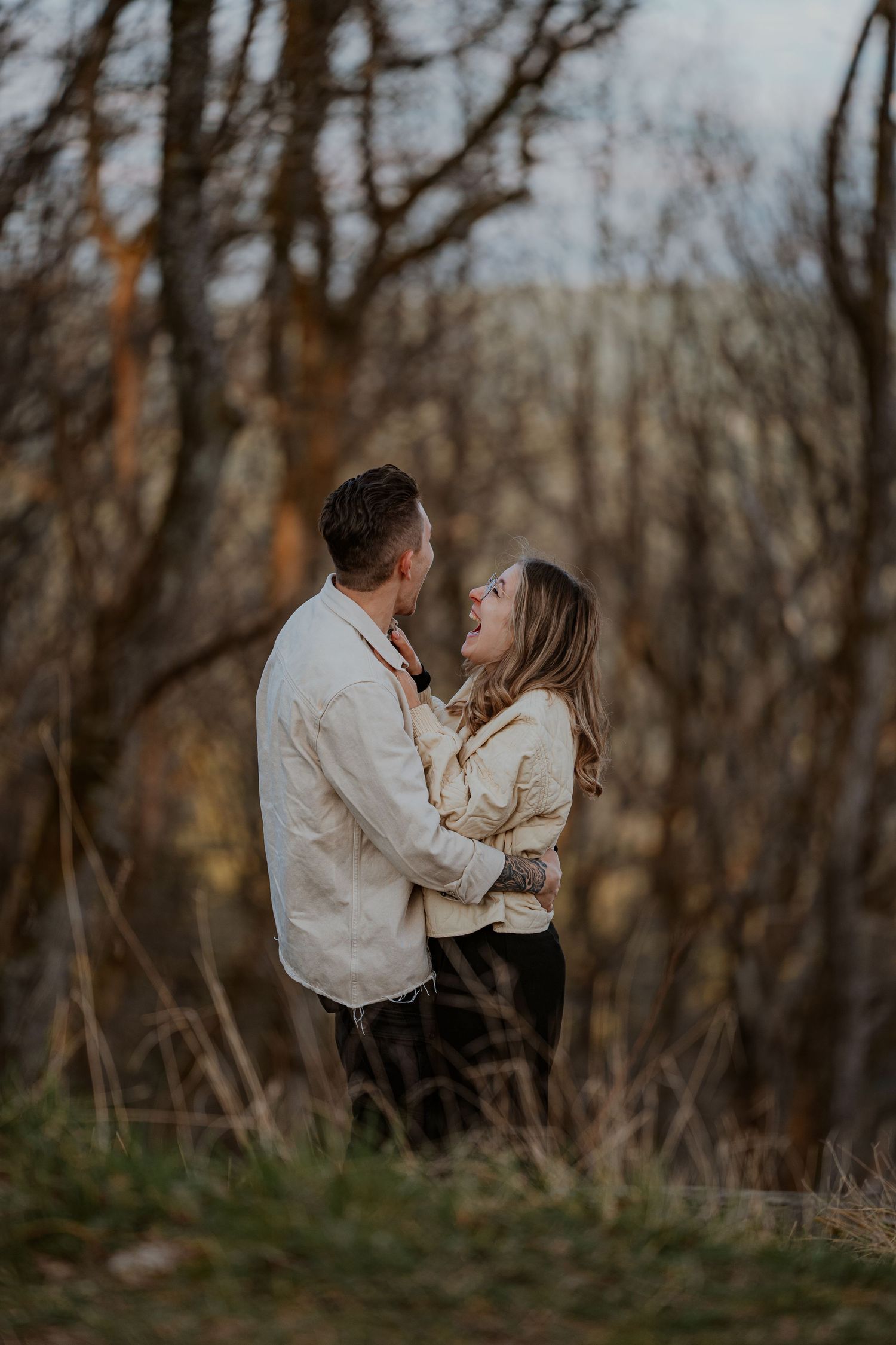 A romantic couple in white clothing shares an intimate moment in a wooded setting during golden hour.