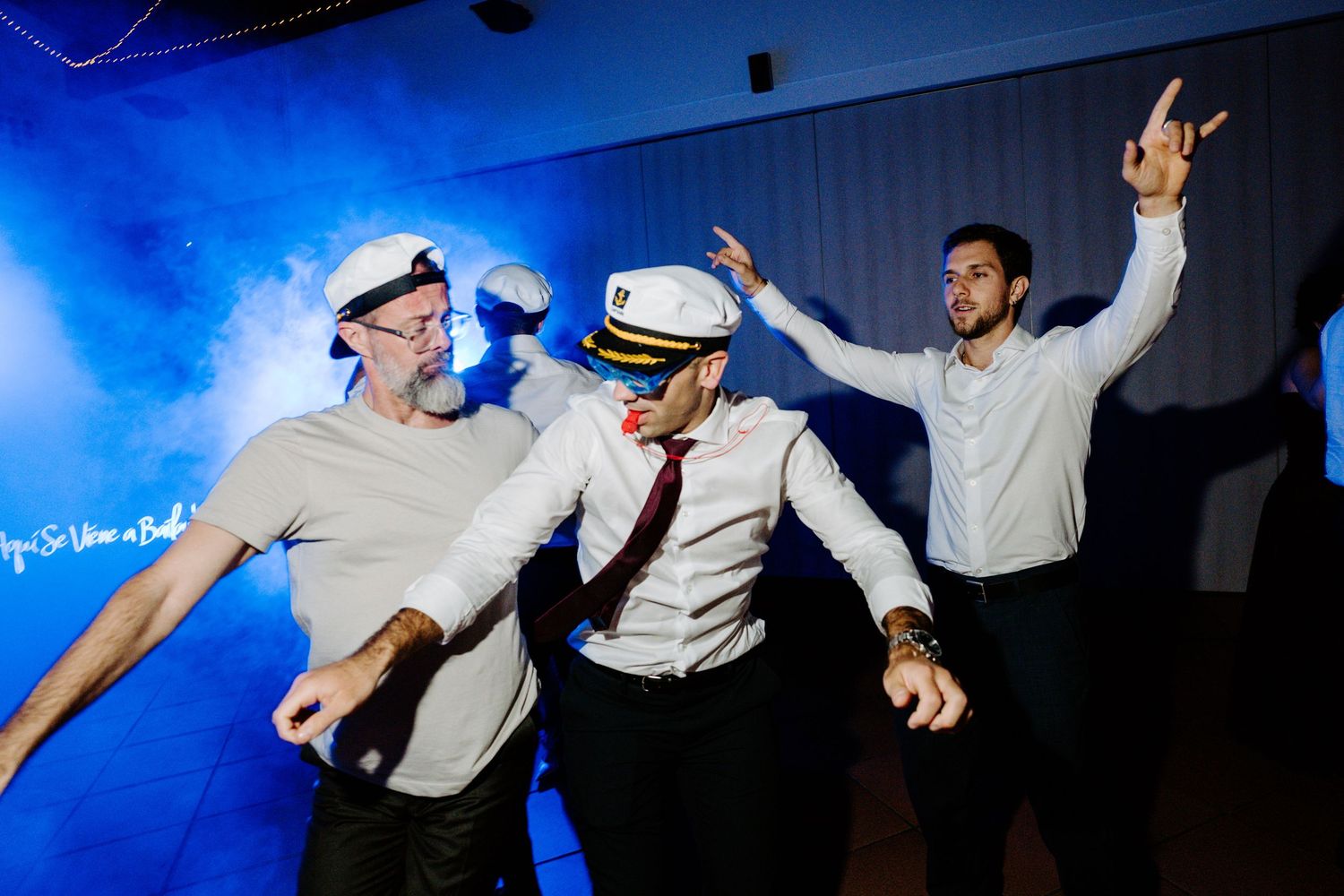 Wedding guests dance energetically under blue lighting at a lively reception celebration.