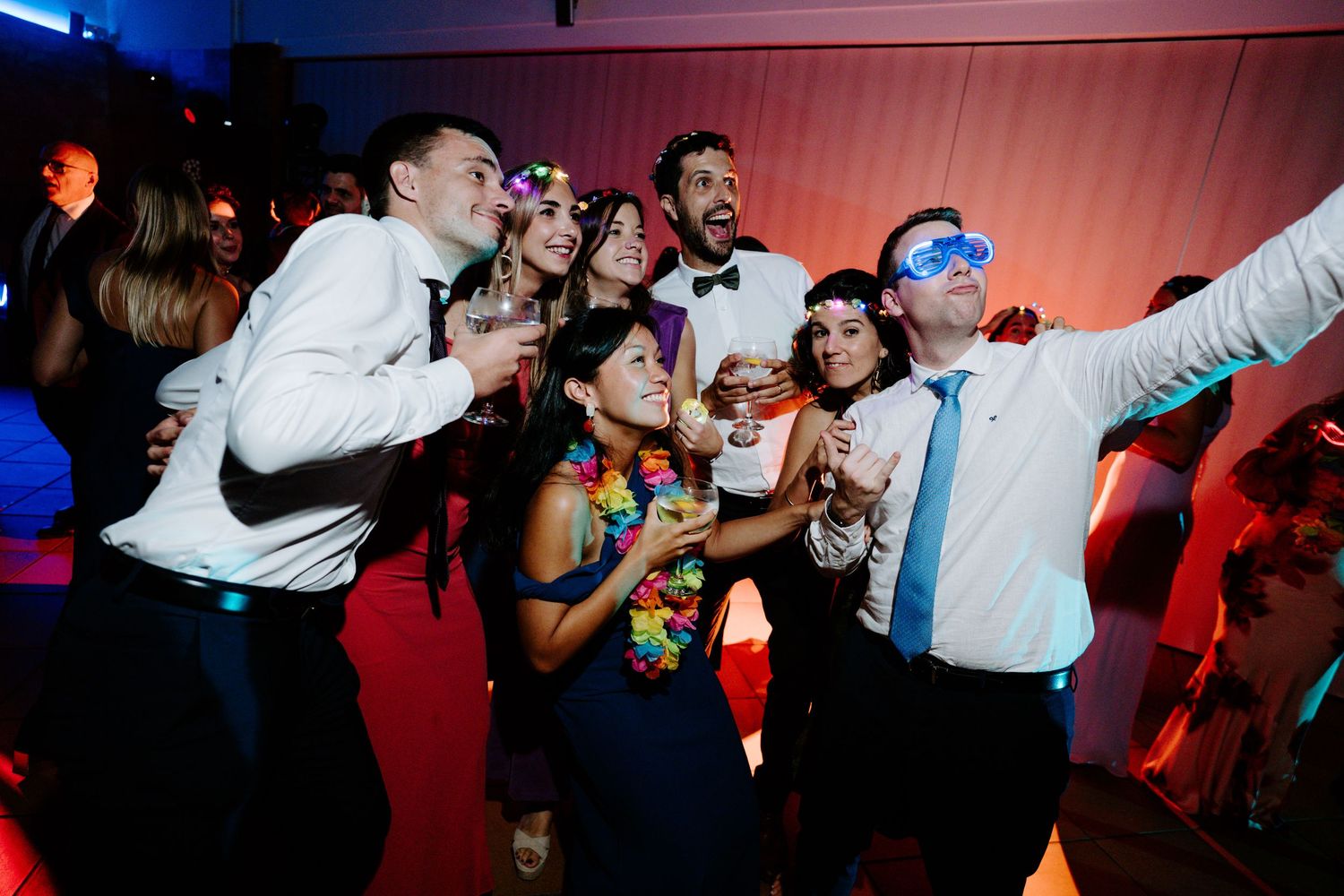 Party guests dancing and celebrating under pink and blue mood lighting at an indoor event.