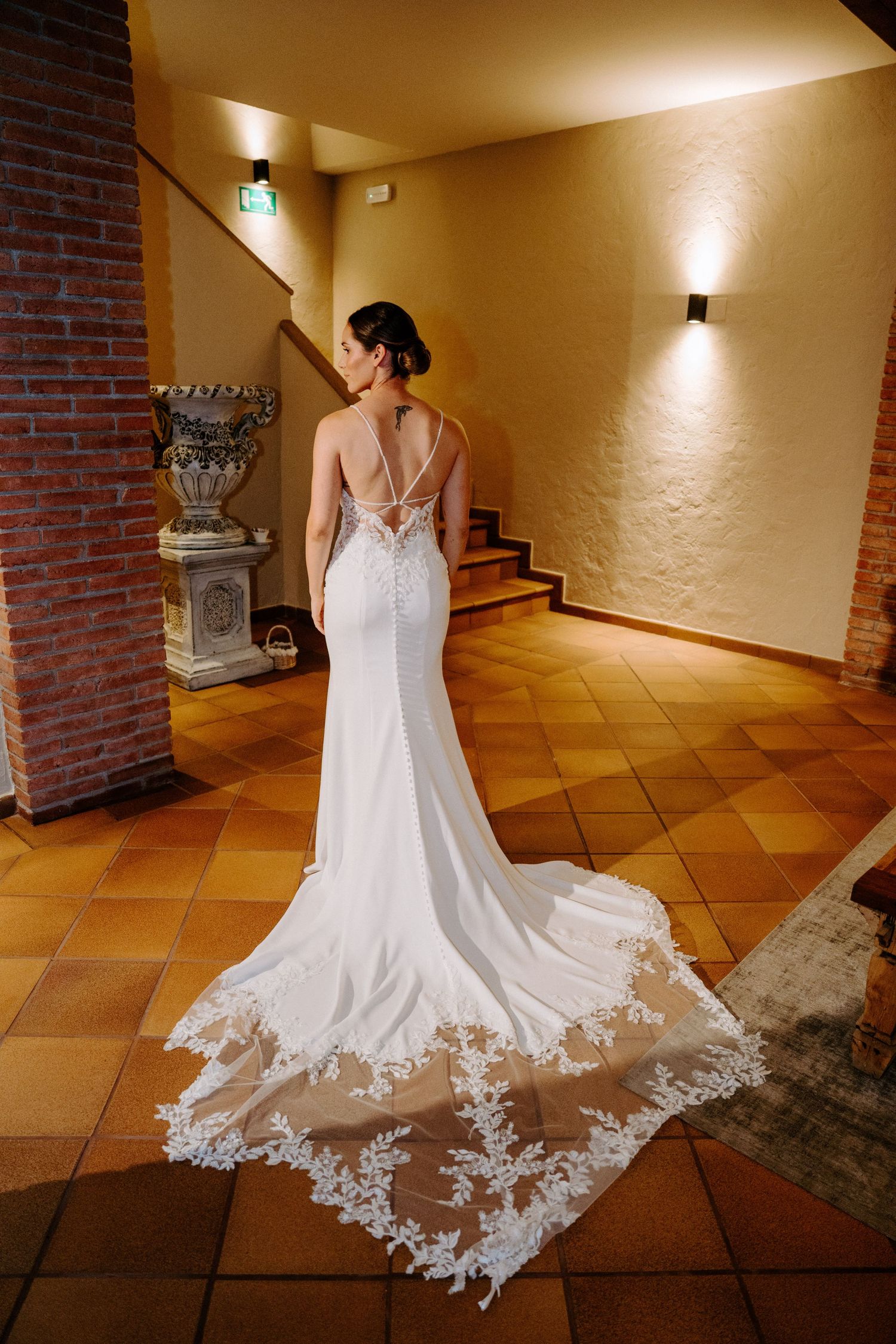 Wedding dress with intricate lace train displayed on terra cotta tile floor against brick wall.
