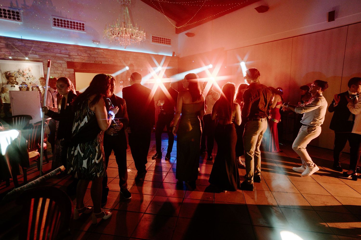 Wide shot of a dimly lit dance floor with party guests celebrating under red ambient lighting.