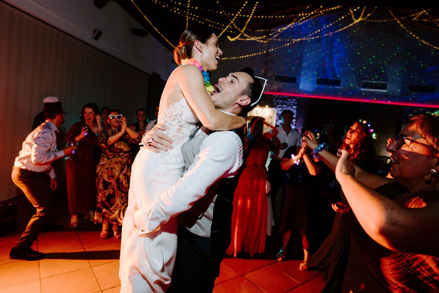Partygoers dancing and celebrating under mixed colored lighting at an indoor venue.