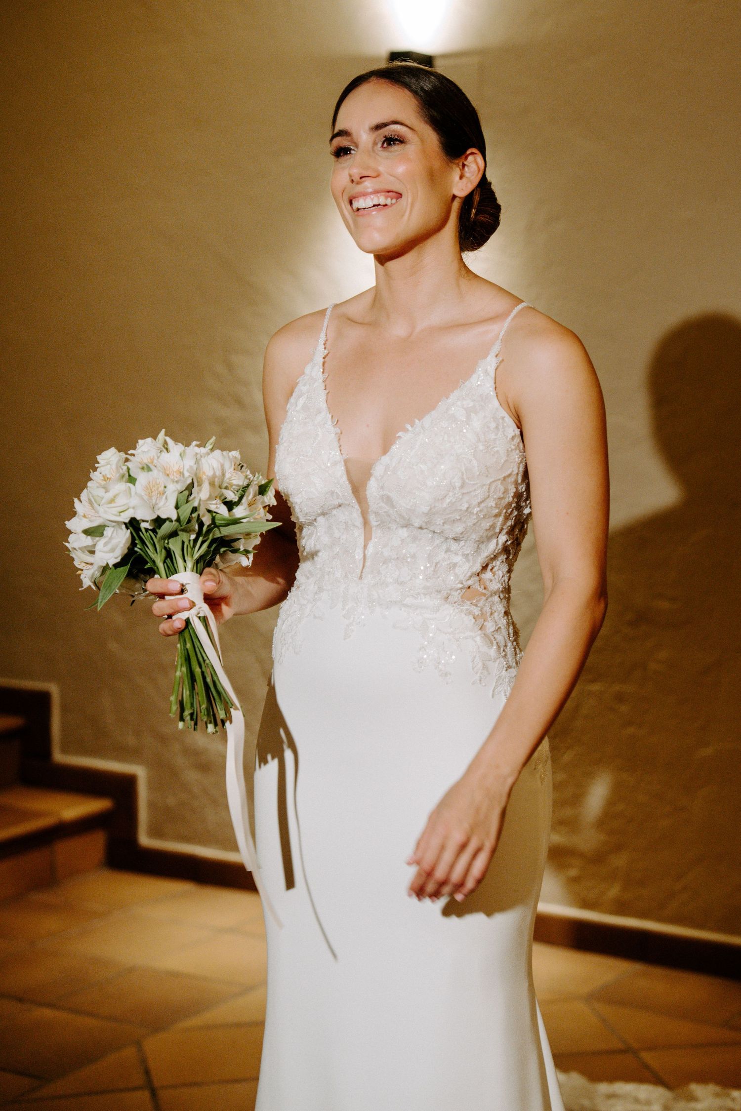 Bride holding white bouquet in elegant lace dress against warm interior backdrop.