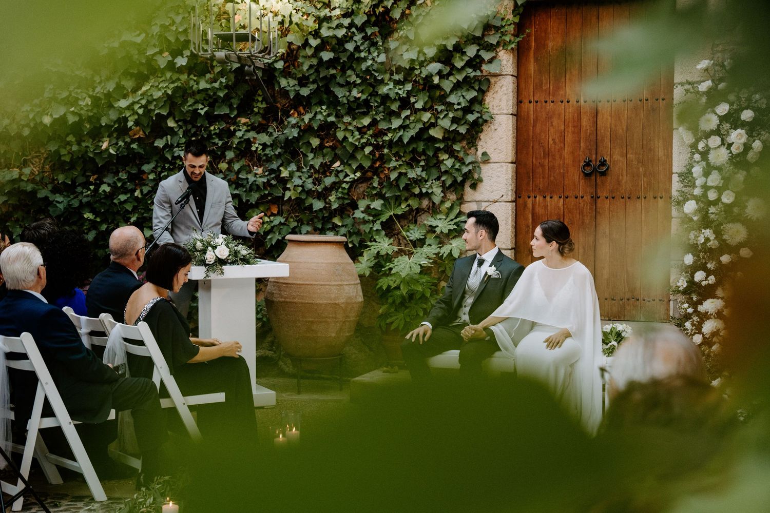 Garden wedding ceremony viewed through greenery with guests seated on white chairs.