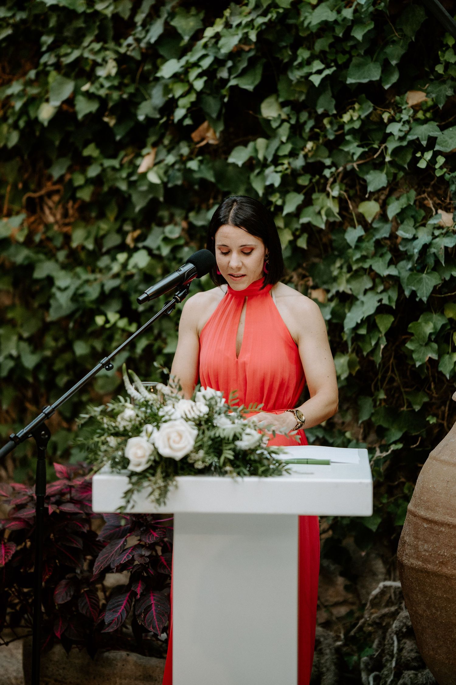 Speaker at white podium against lush green ivy backdrop during outdoor ceremony.