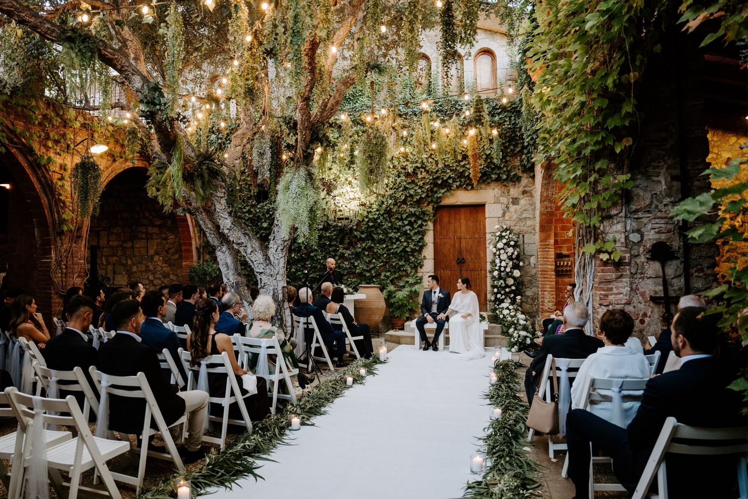 An outdoor wedding ceremony setup with white chairs and string lights in a rustic courtyard with ivy-covered walls.