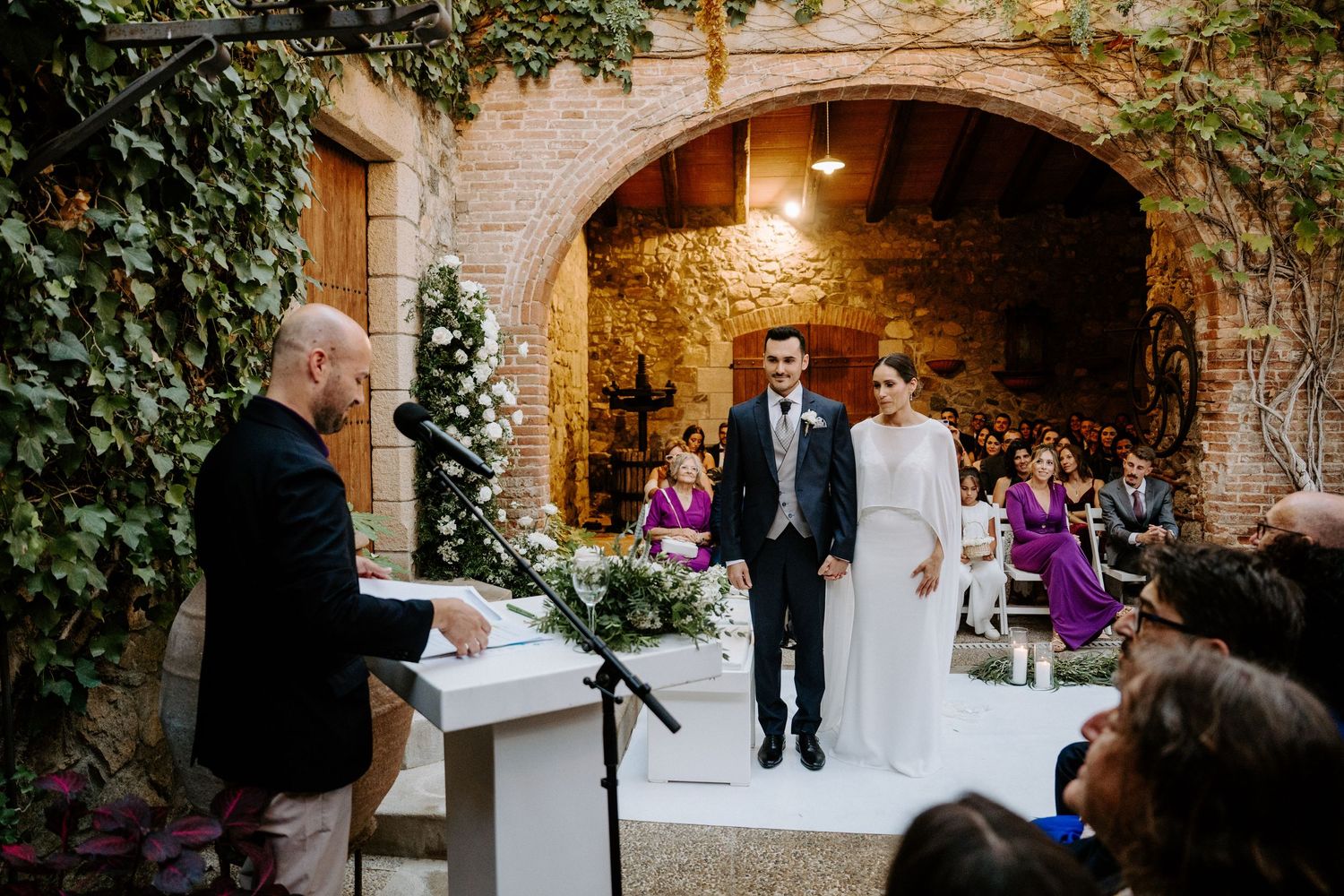 A wedding ceremony taking place in a stone courtyard with ivy-covered walls and white floral arrangements.