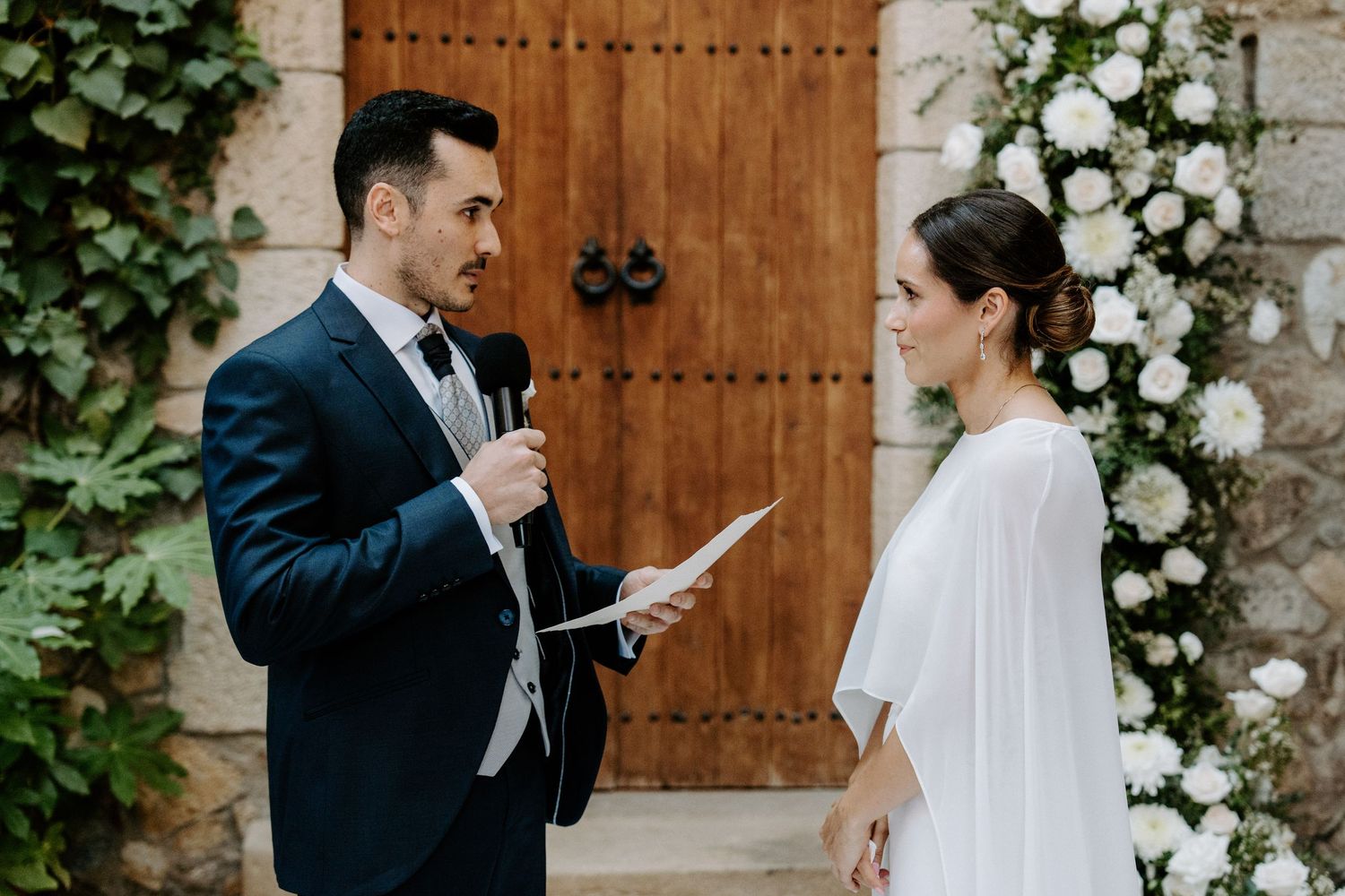A wedding couple exchanging vows in front of a rustic wooden door with white floral arrangements.