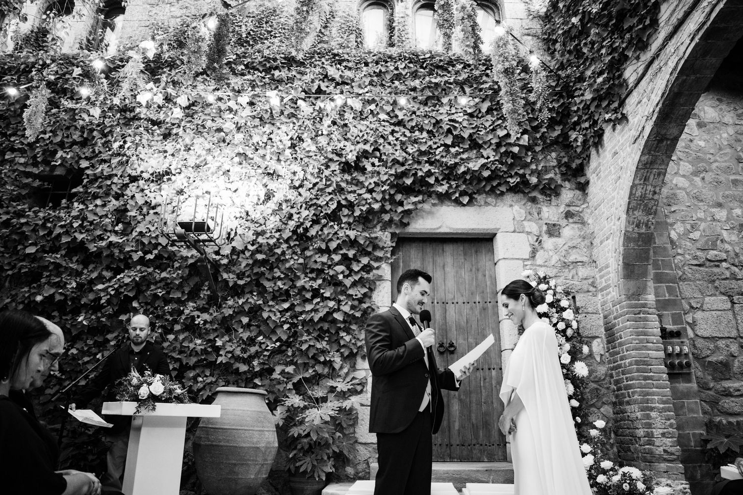 A black and white photo of a wedding ceremony in a stone courtyard with climbing ivy.