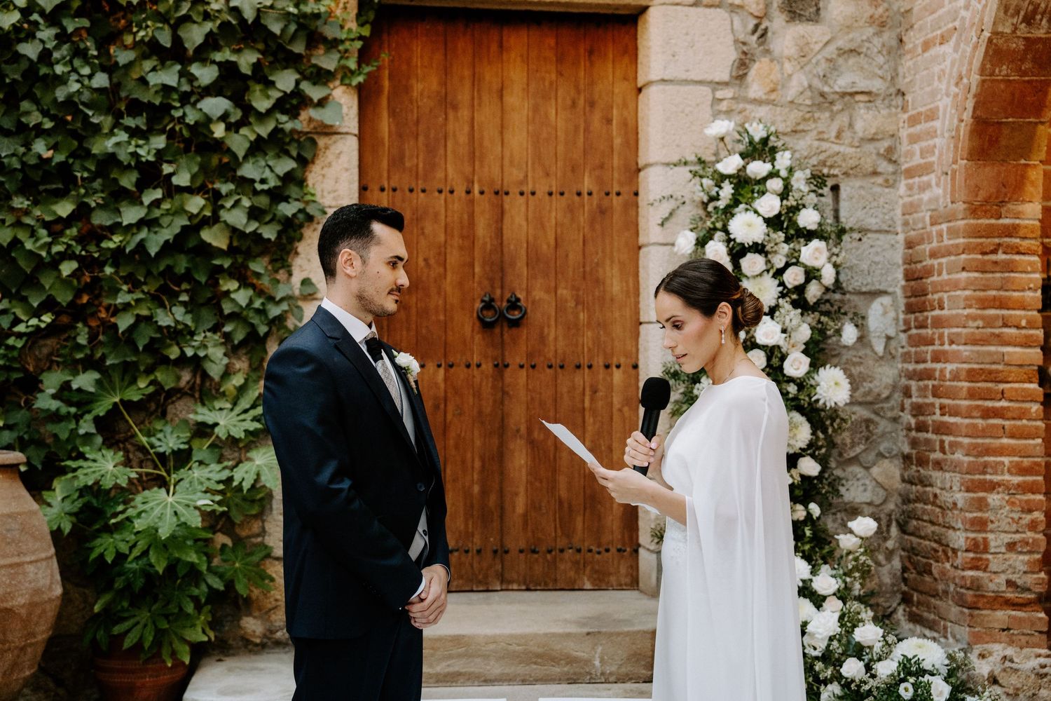 A couple exchanging vows in front of a wooden door decorated with white flowers and greenery.
