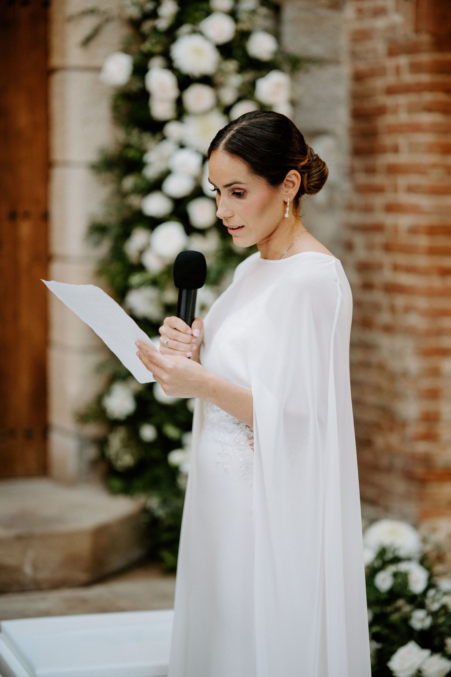 A bride in an elegant white cape dress reading from a paper during the ceremony.