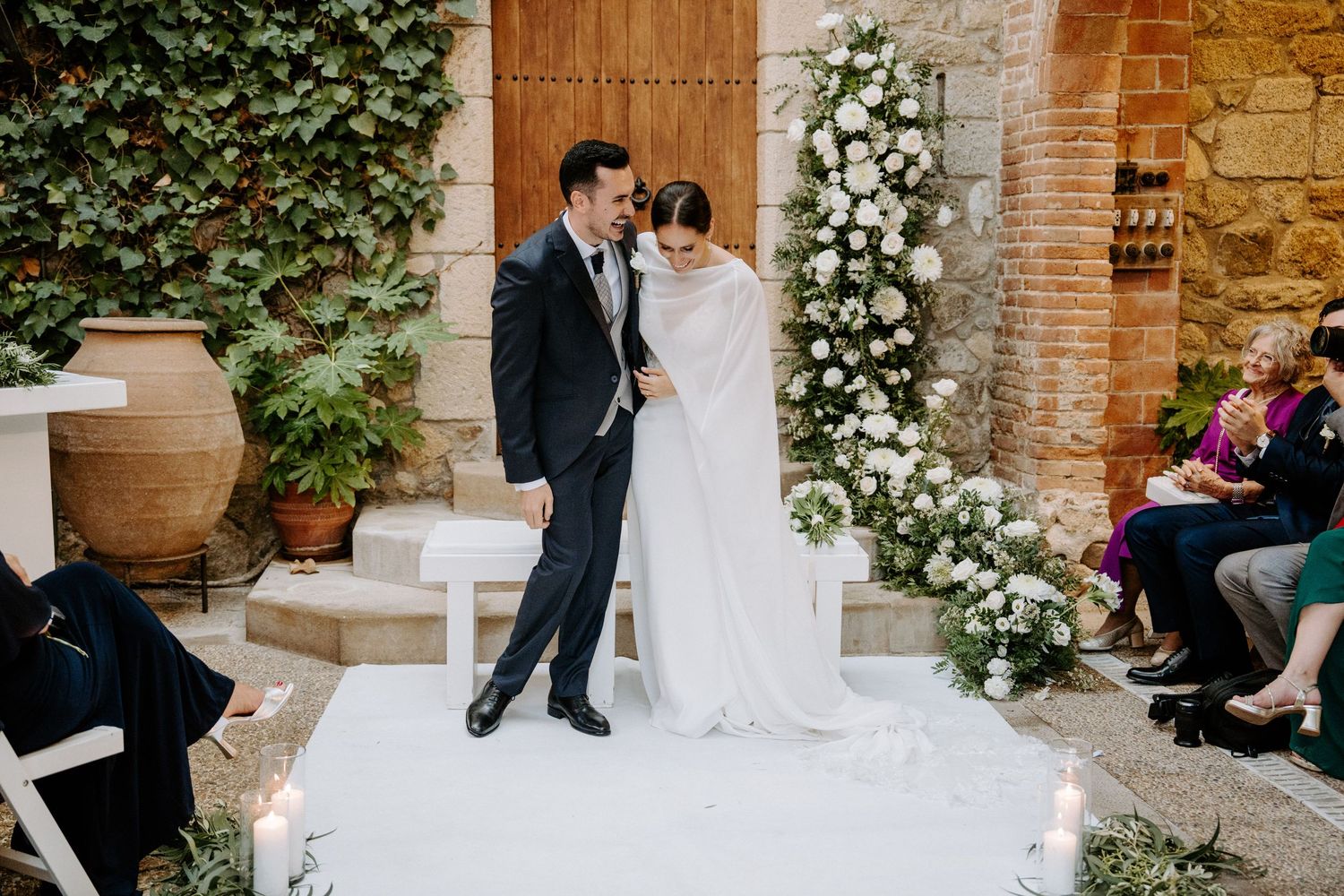 Ceremonia de boda con sillas blancas y arreglos florales sobre fondo de piedra y hiedra. Mas de Sant Llei