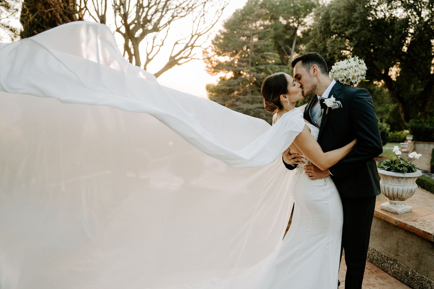 A flowing white wedding veil catches the golden sunset light as a couple shares a romantic moment outdoors.