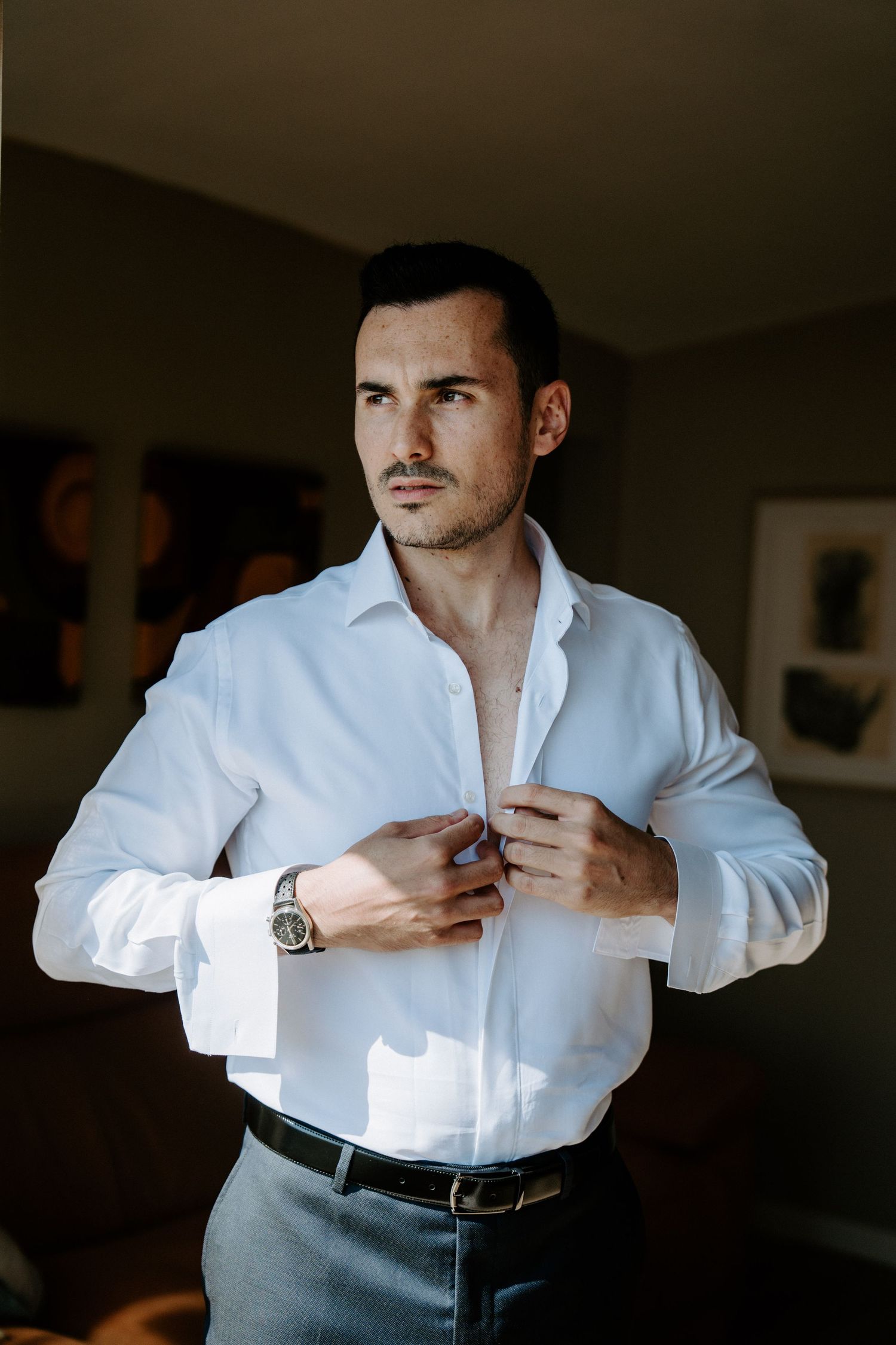 Close-up shots of groom buttoning white dress shirt in moody lighting.
