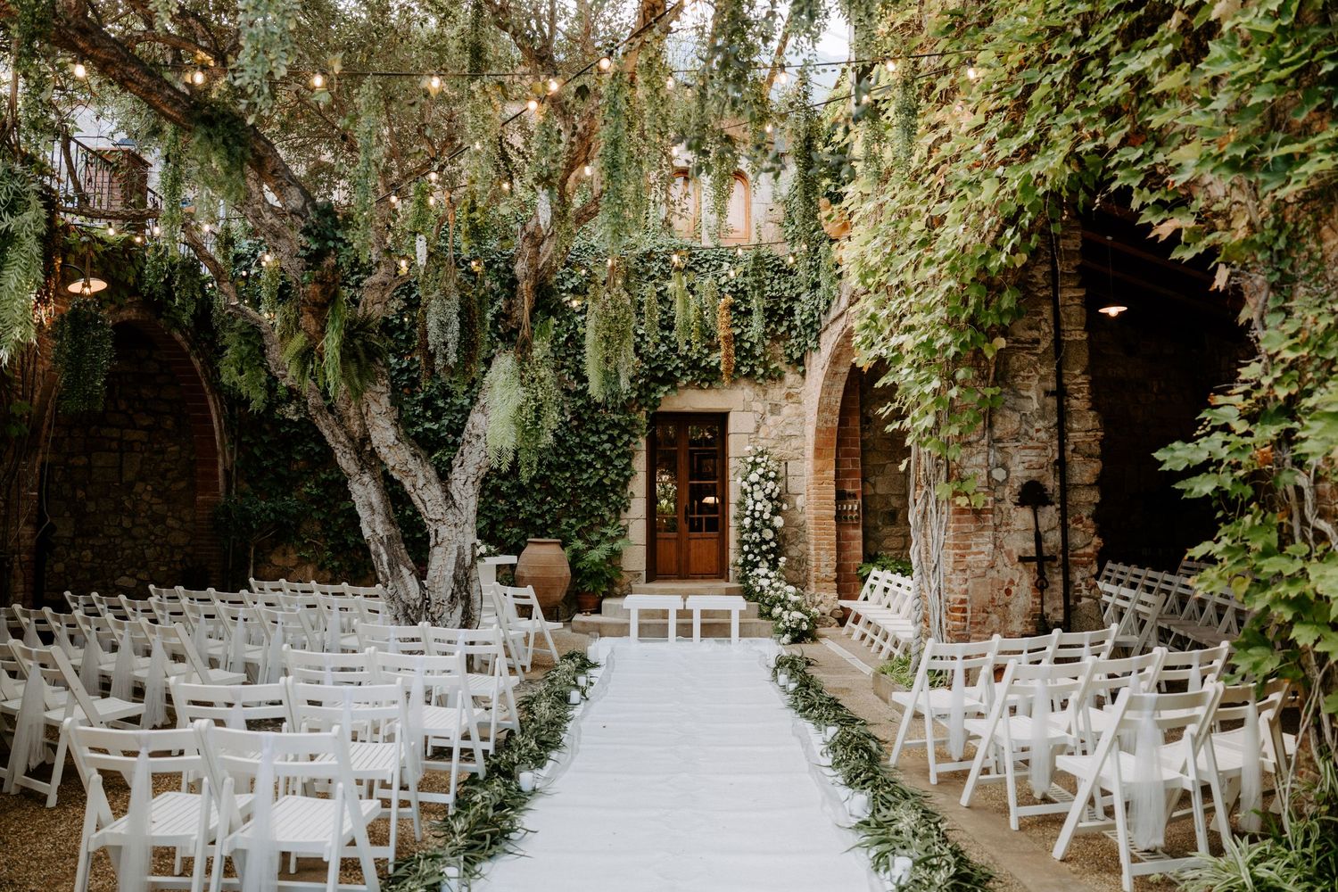 Outdoor wedding ceremony setup with white chairs under ivy-covered trees.