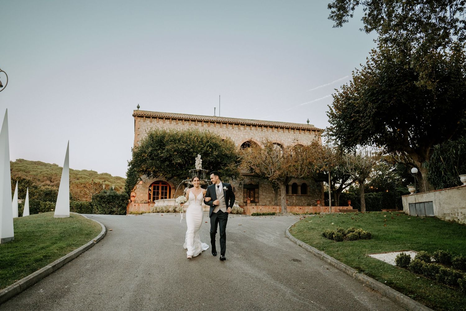 Wedding couple walking down driveway of Mediterranean style villa with mountain backdrop.
