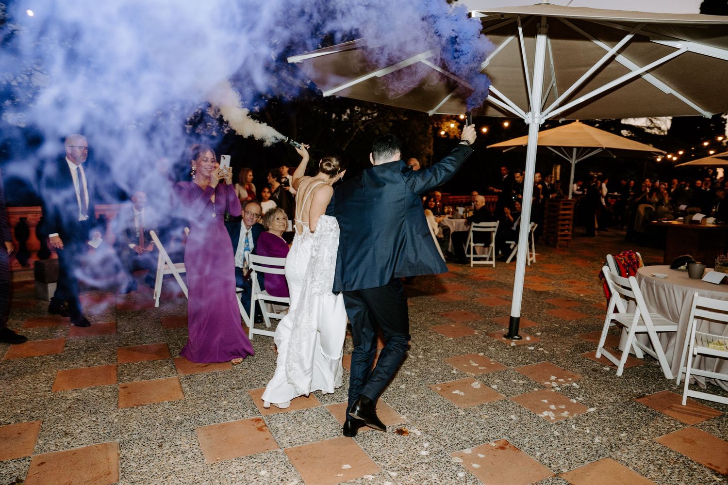 Wedding party dancing outdoors under white umbrellas with blue smoke effects.
