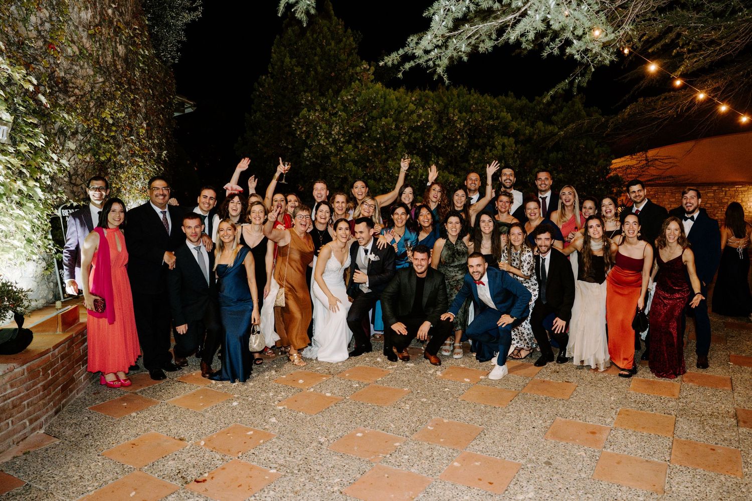 Large group photo of wedding guests gathered outdoors under string lights at night.