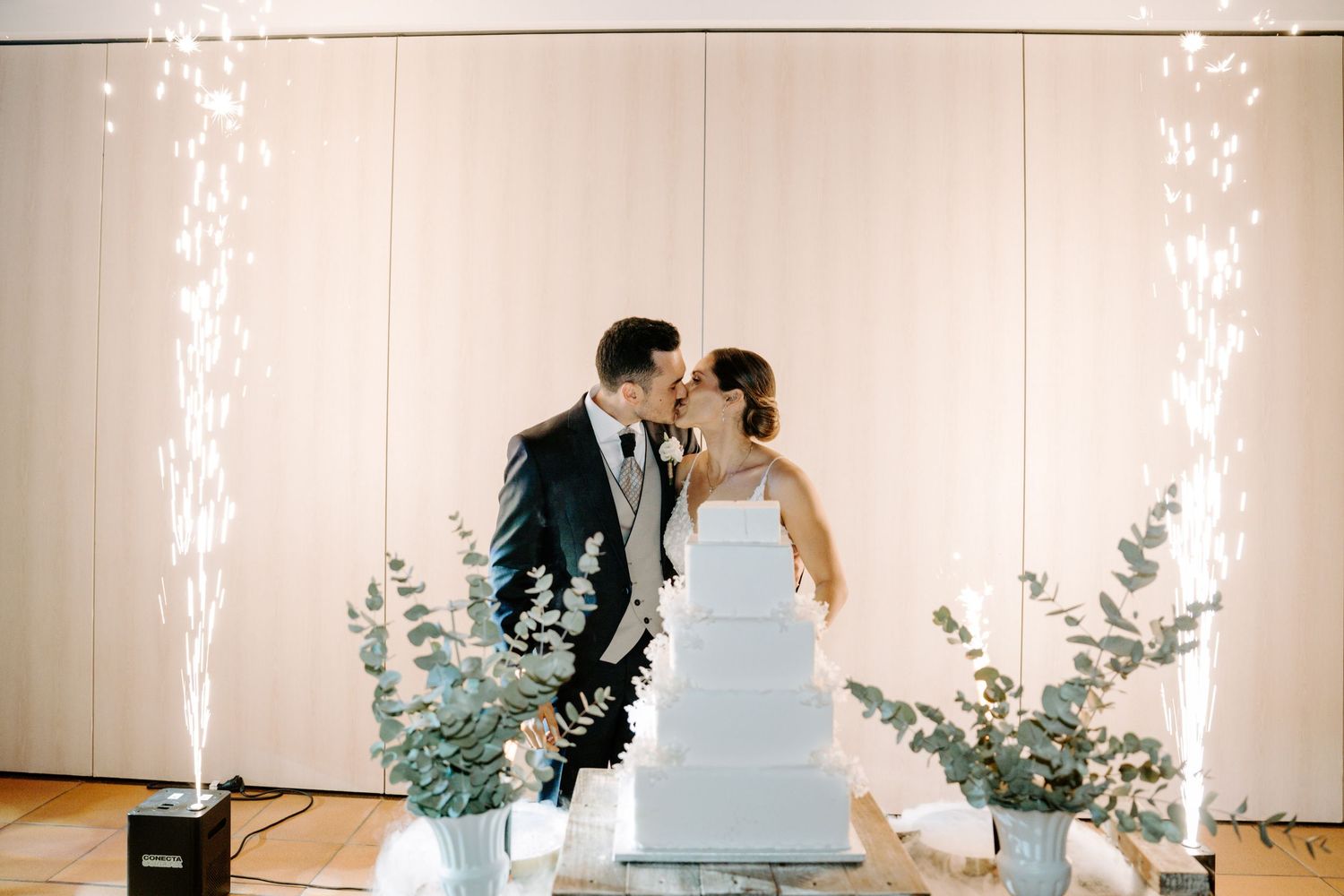Elegant wedding reception with couple sharing a romantic moment near a white tiered cake and floral decorations.