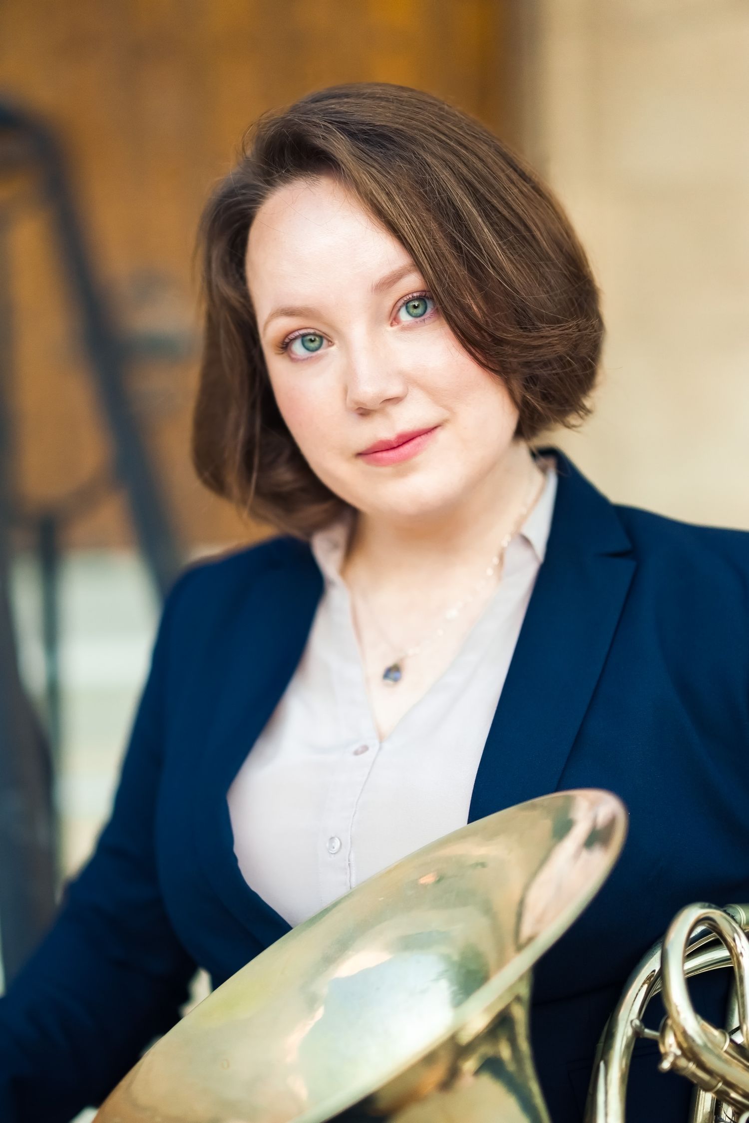 A woman with short brown hair hold a French horn, sitting on the steps of a church.