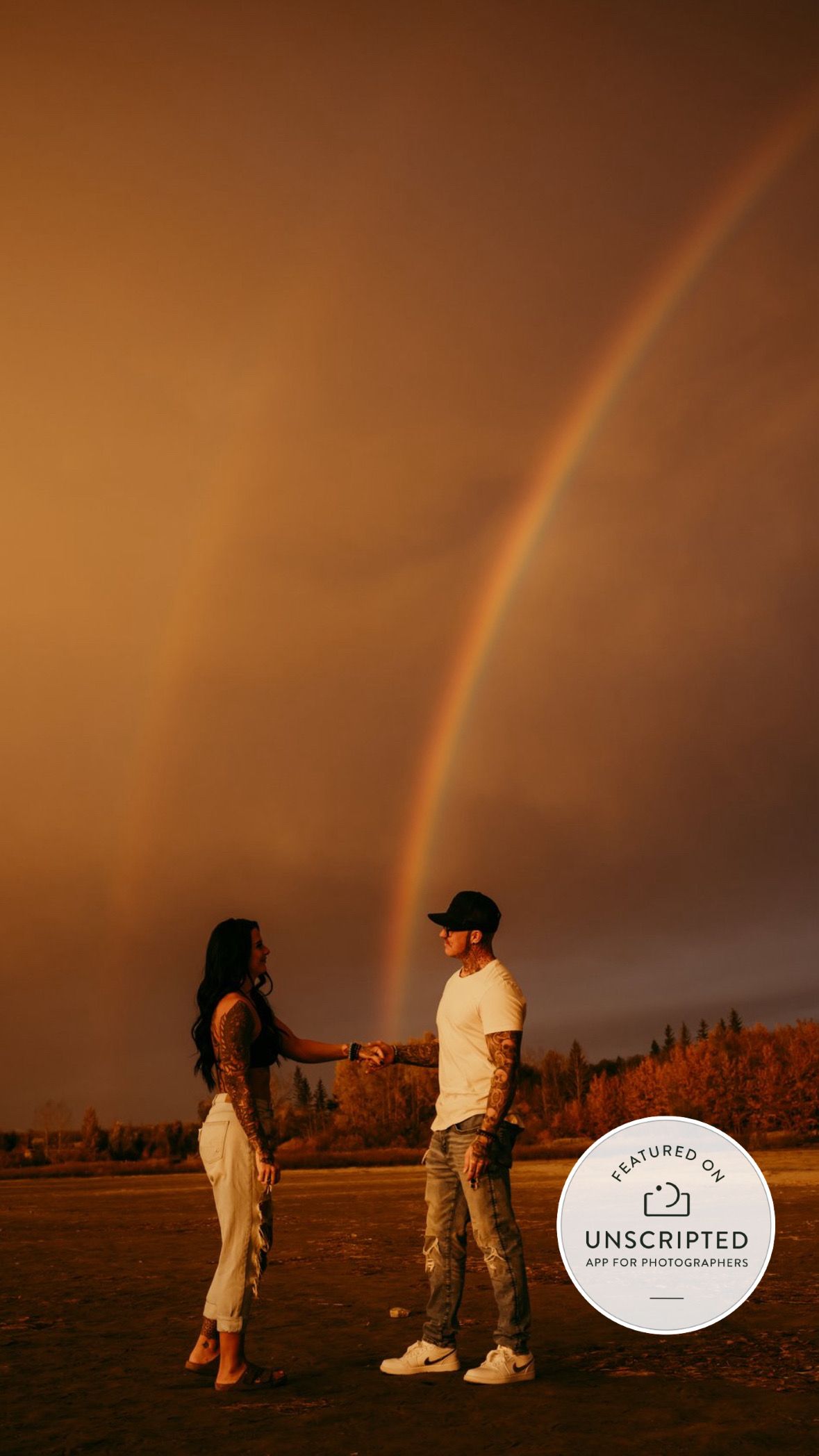 Couple holds hands against dramatic desert sunset sky with double rainbow and saguaro cacti silhouettes in background.
