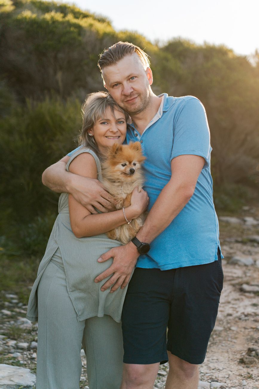 During a maternity photoshoot in Sydney a couple hugs outdoors at sunset with their small Pomeranian dog while expecting a baby