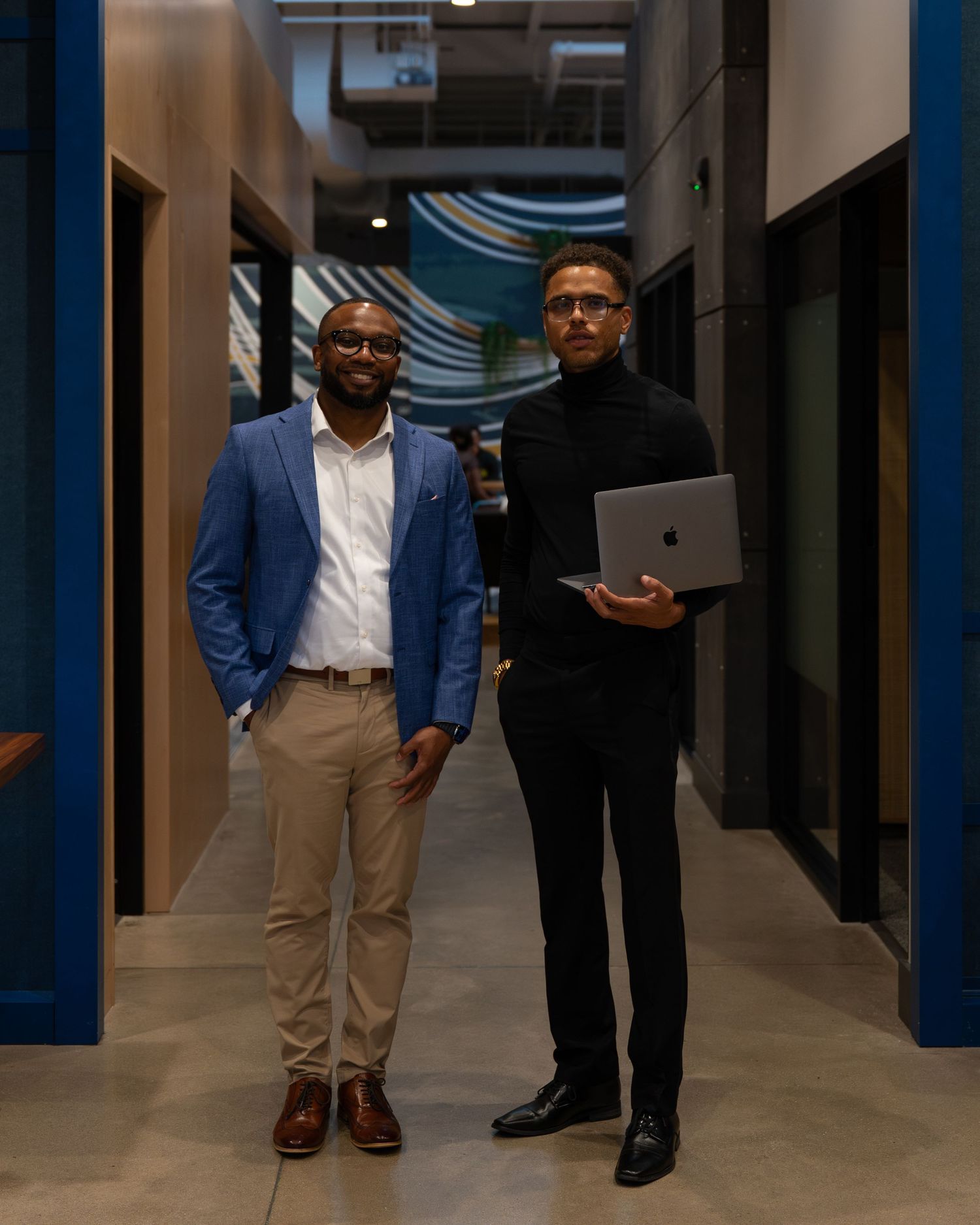 Two businessmen in professional attire walk down a modern office hallway discussing work.