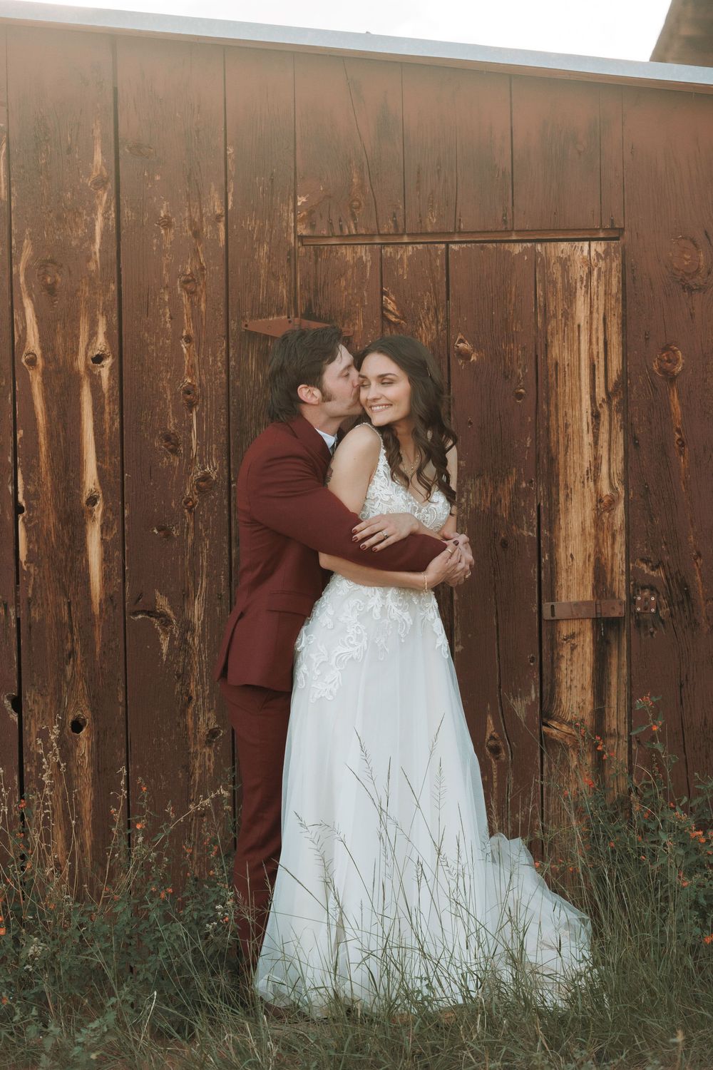 Groom in burgundy suit kissing bride in lace wedding dress beside rustic wooden barn.