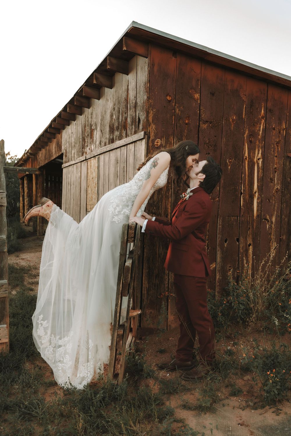 Abiquiu Groom lifts bride in a romantic dip kiss against a rustic red wooden barn at their outdoor wedding at Ghost Ranch.