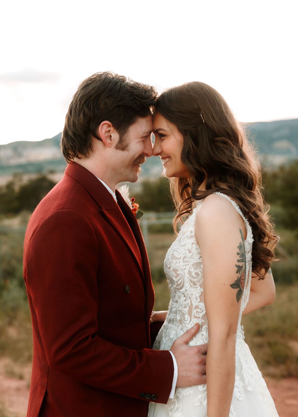 Smiling bride and groom in burgundy suit share a tender forehead-touch moment in a desert landscape.