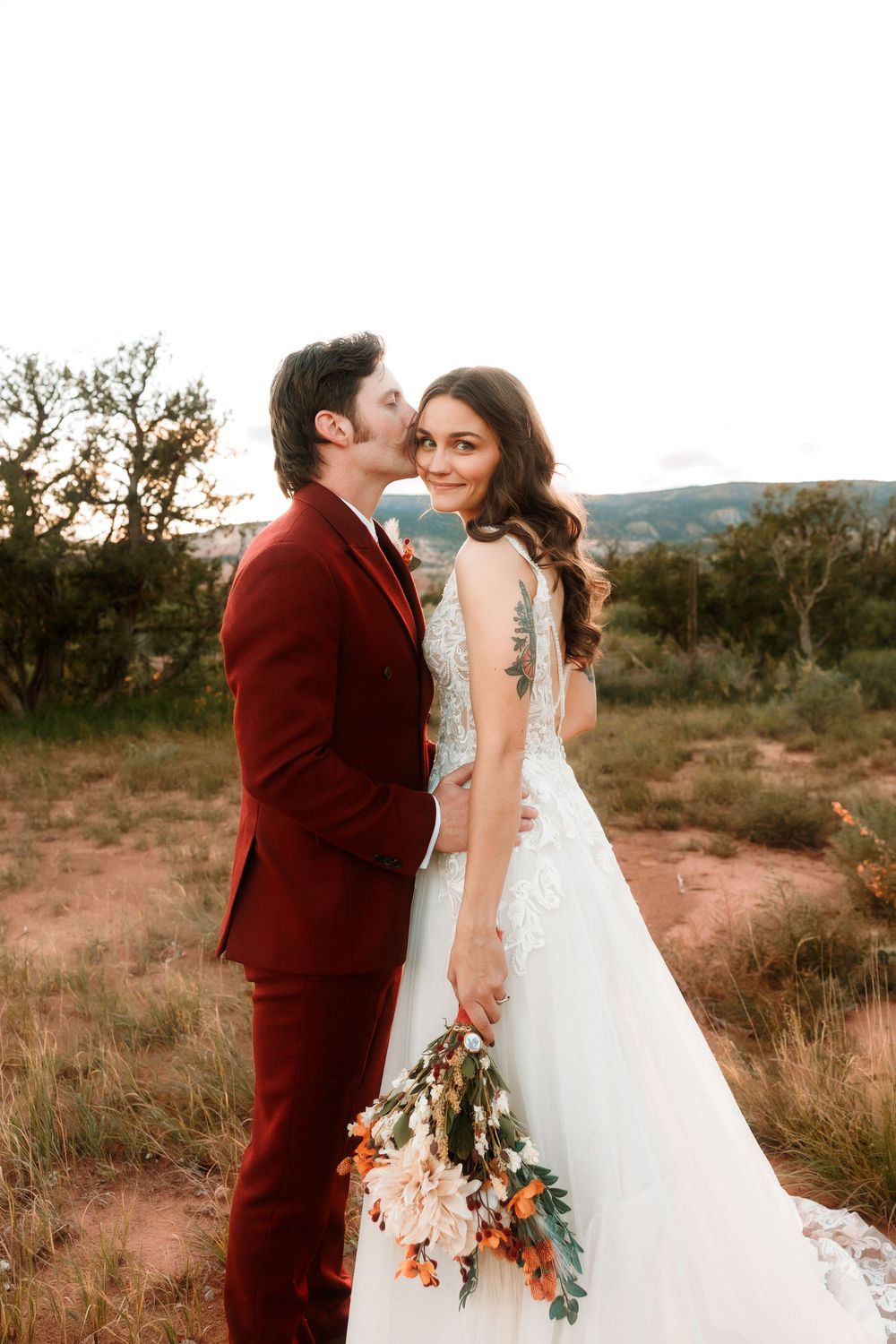 Groom in burgundy suit kisses bride on cheek as she holds a pastel wildflower bouquet in a desert setting.