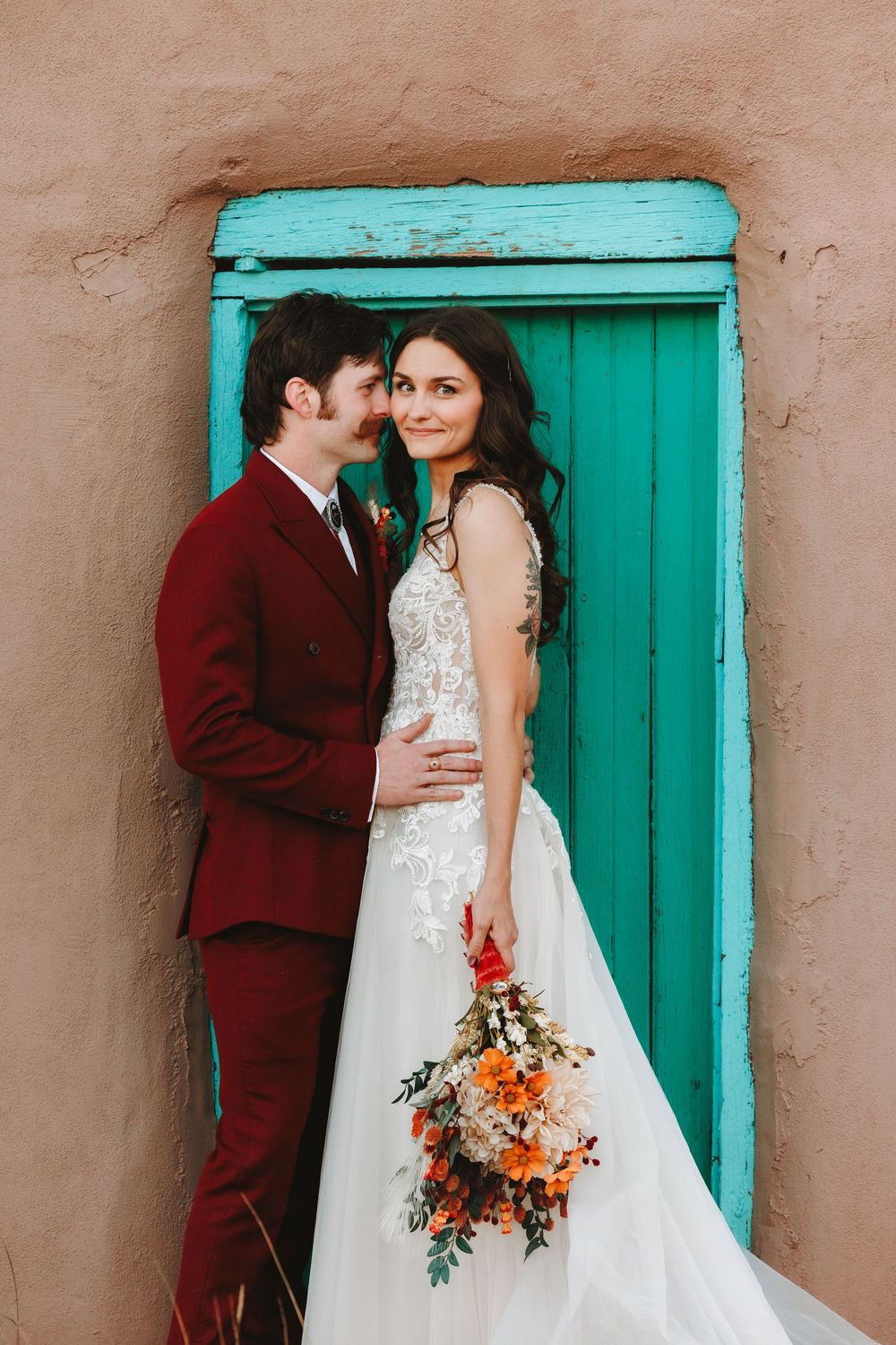 Bride and groom pose before a turquoise door on an adobe wall, she holds an orange and red bouquet.