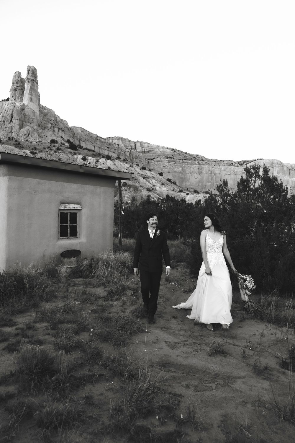 Black and white photo of bride and groom walking near an adobe building with dramatic rock formations behind.