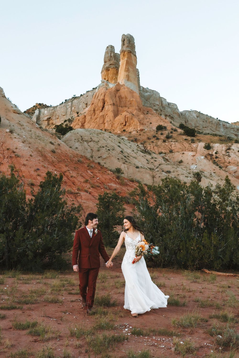 Bride and groom hold hands walking through red desert landscape with towering sandstone rock formations above.
