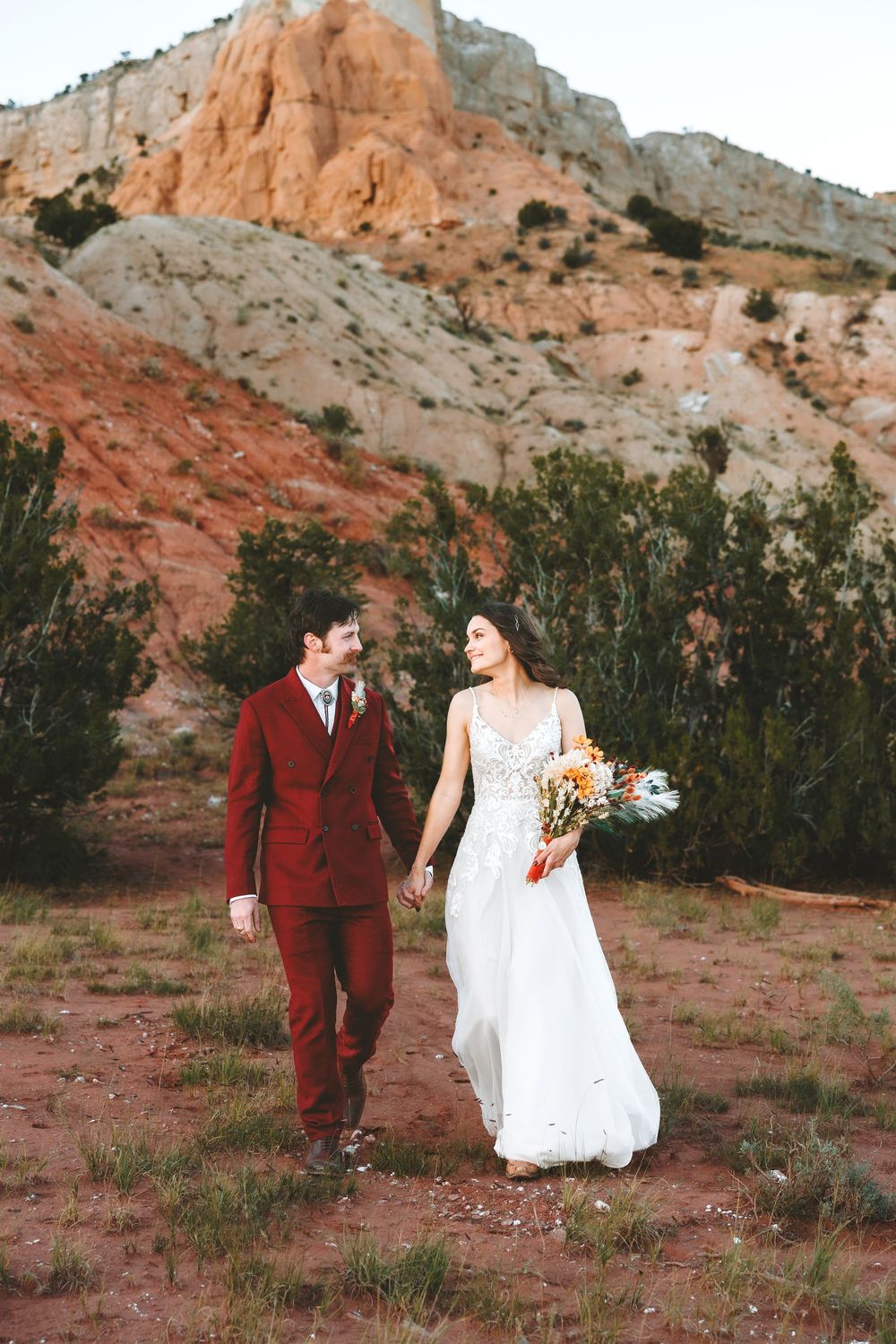 Couple in wedding attire walks hand in hand through a red rock desert landscape at golden hour.