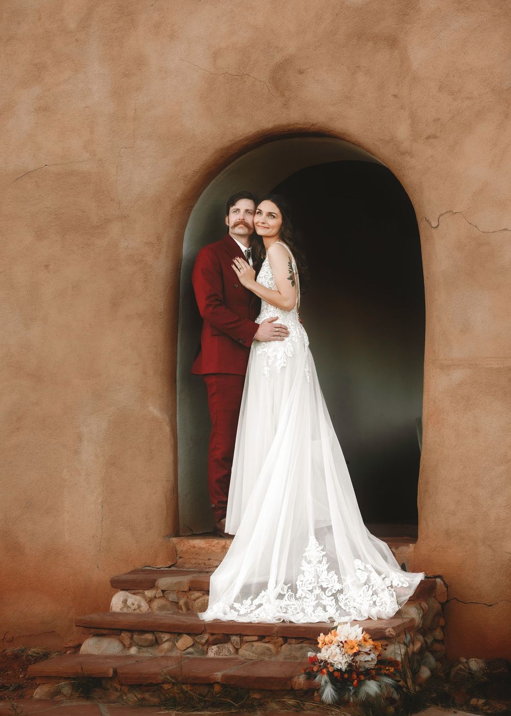 Bride and groom embrace in an arched adobe doorway, her lace gown trailing down stone steps below.