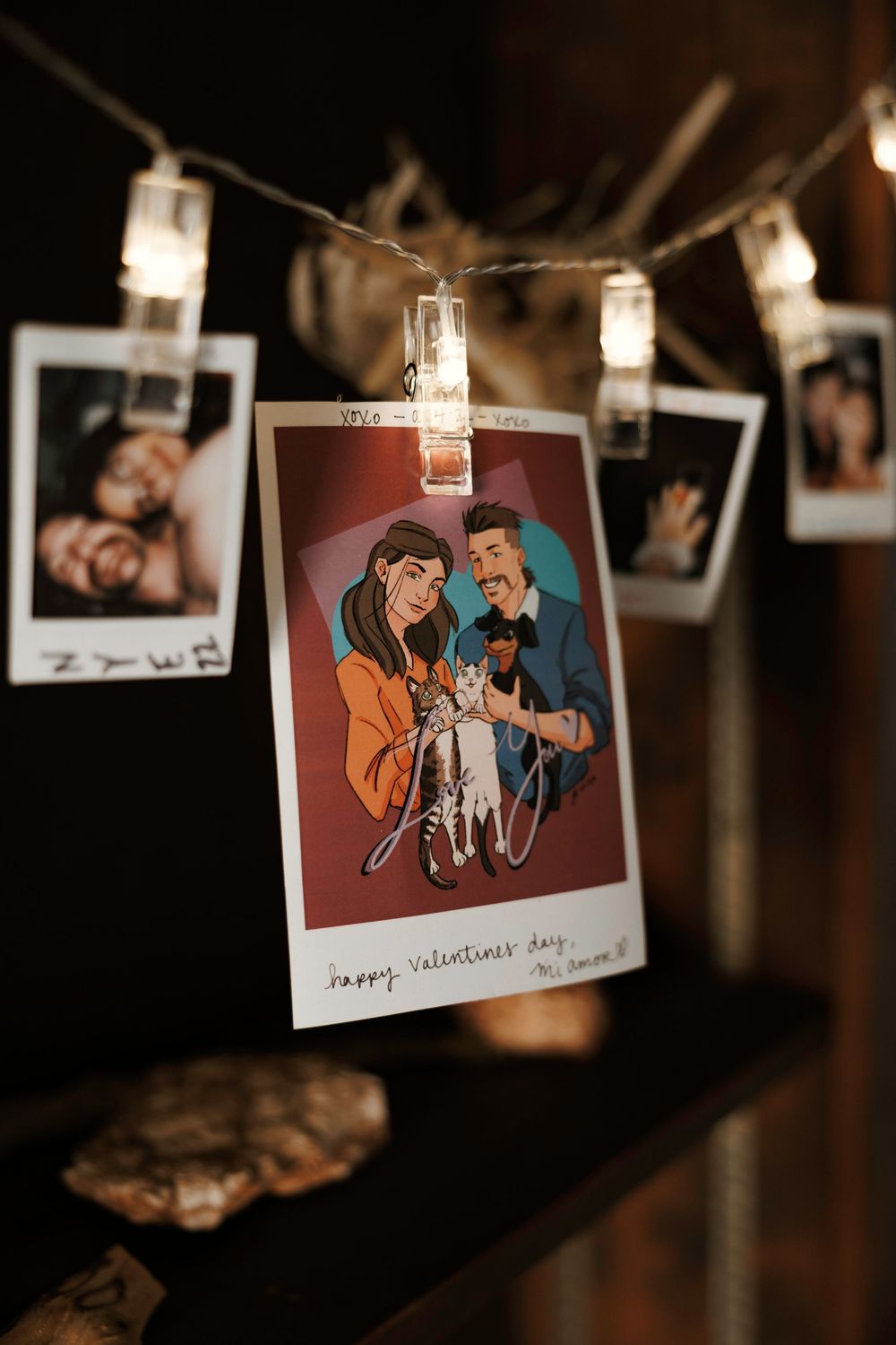Illustrated Valentine card and Polaroid photos hang on a string of lights against a dark background.