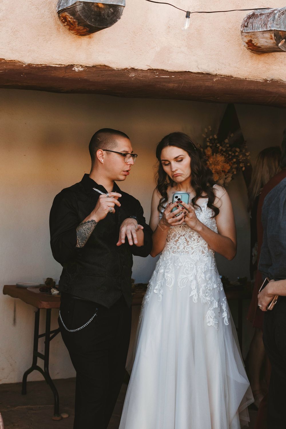 Bride in lace gown and groom in black suit look at a smartphone together at a rustic wedding venue.
