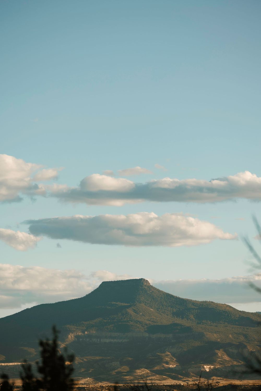 A flat-topped mesa rises against a pale blue sky with scattered white clouds in the Southwest.
