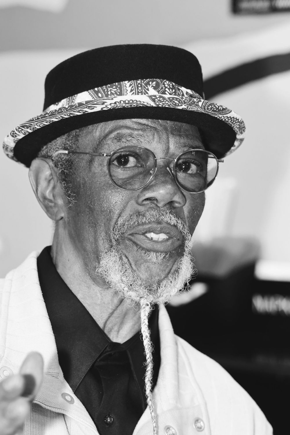 A black and white portrait of an older man wearing glasses, a black hat with decorative band, and white jacket.