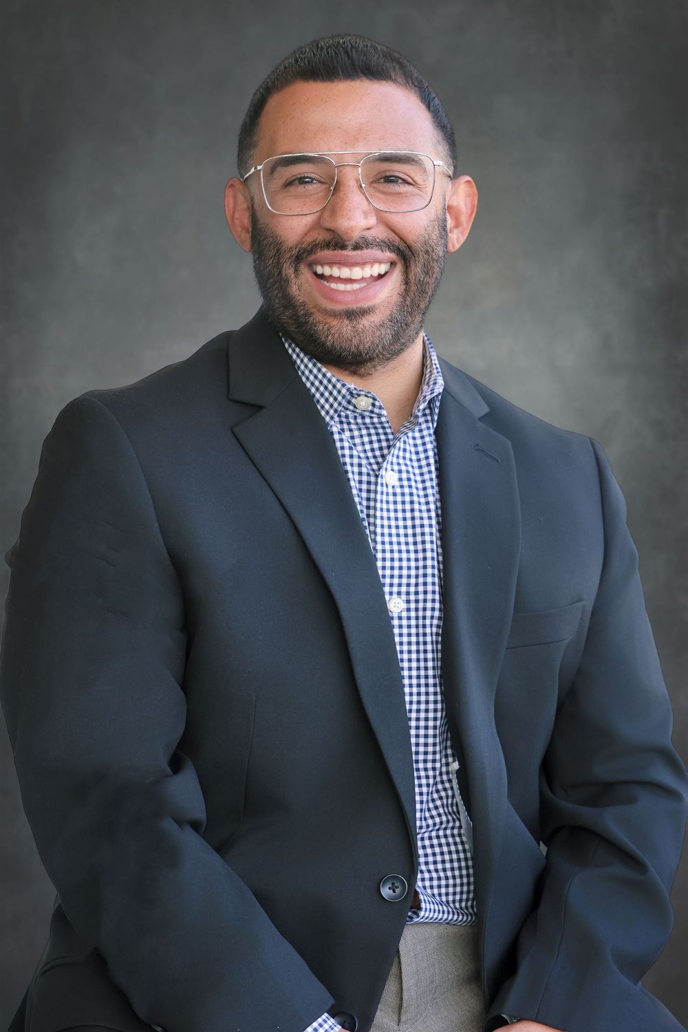 Professional headshot of person in checkered shirt and blazer with glasses.