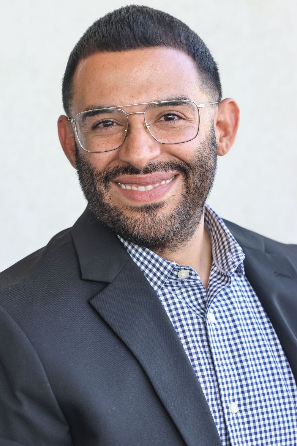 Professional headshots showing warm smile wearing checkered shirt and dark blazer against light background.