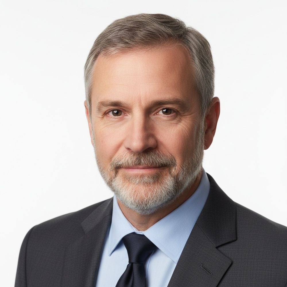 Professional headshot of person in dark suit and blue tie with gray hair and beard against white background.