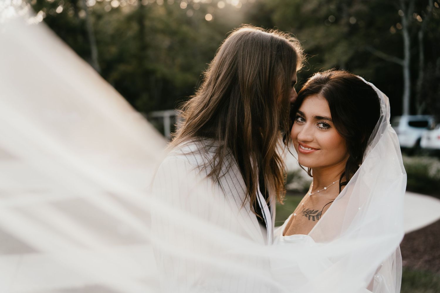 A bride wearing a flowing white veil stands outdoors during a wedding photoshoot at sunset.