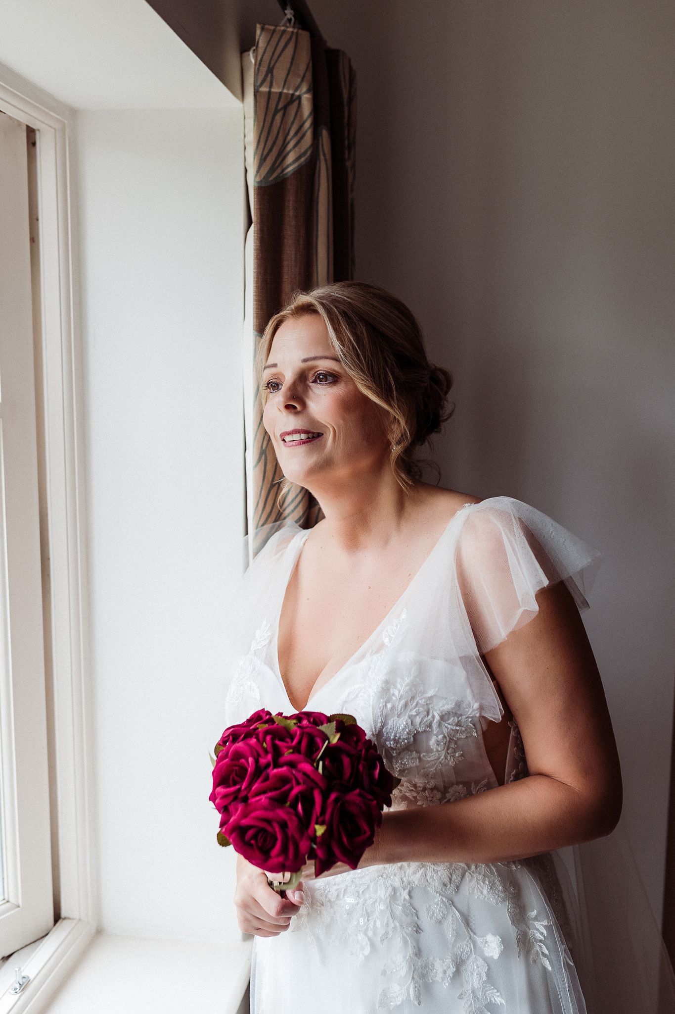 Bride in white lace dress holds deep red rose bouquet while standing by window.