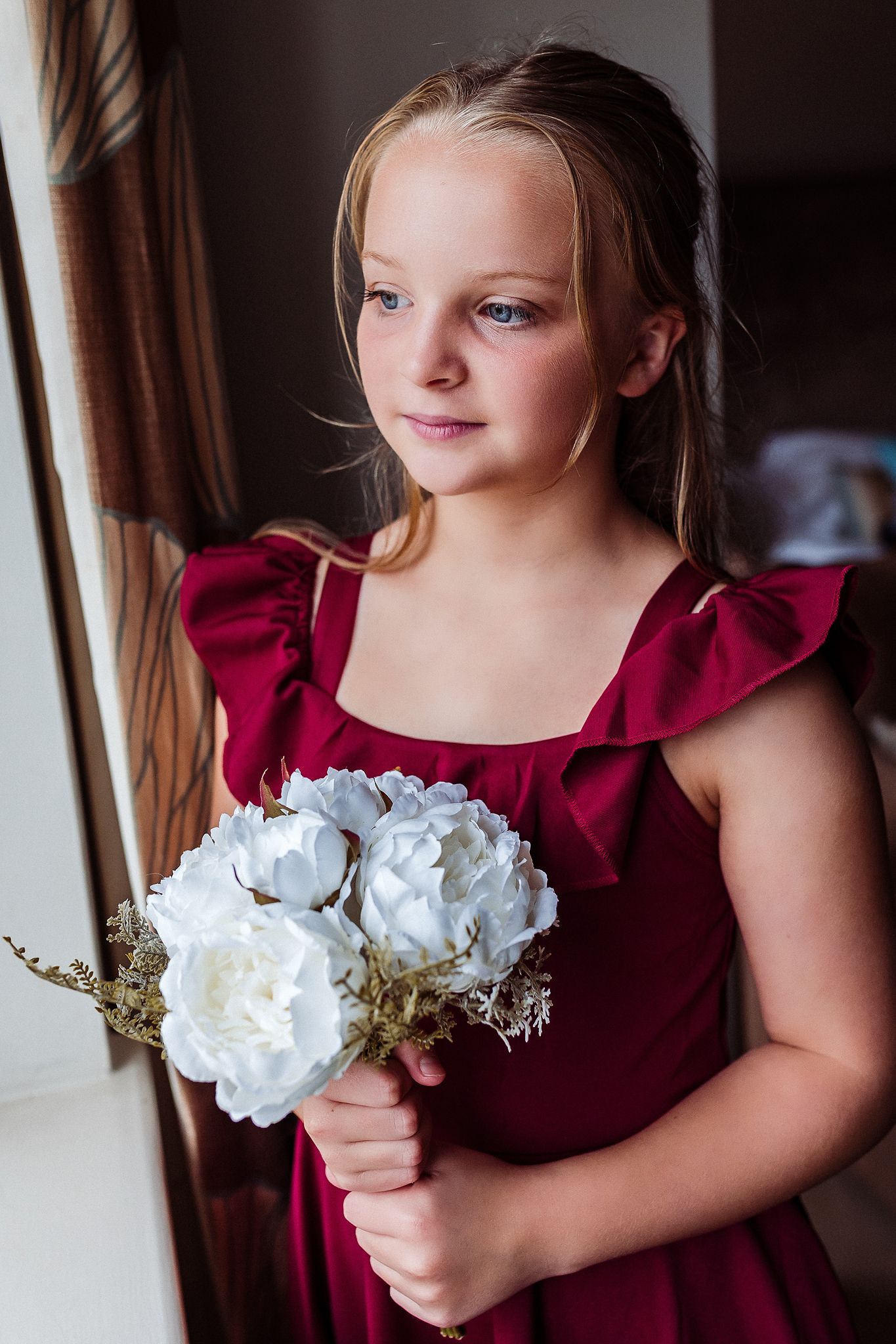 A young attendant in a burgundy dress holds white flowers while standing near a window.