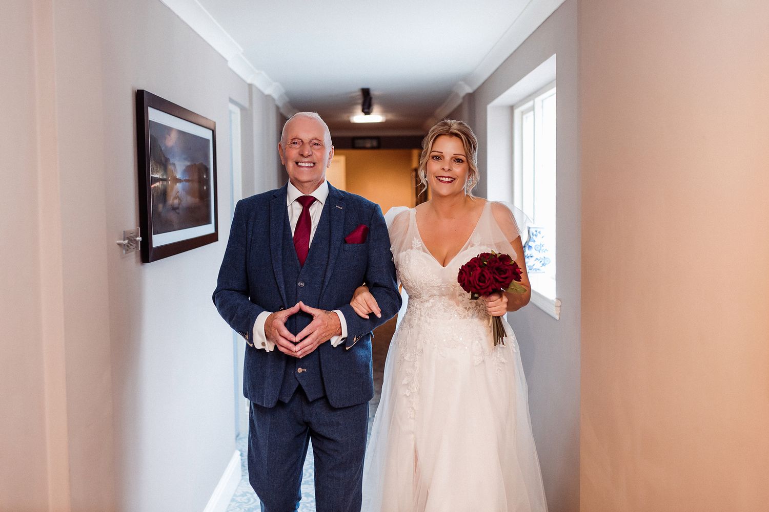 A wedding party poses in formal attire in a bright hallway with natural lighting streaming through windows.