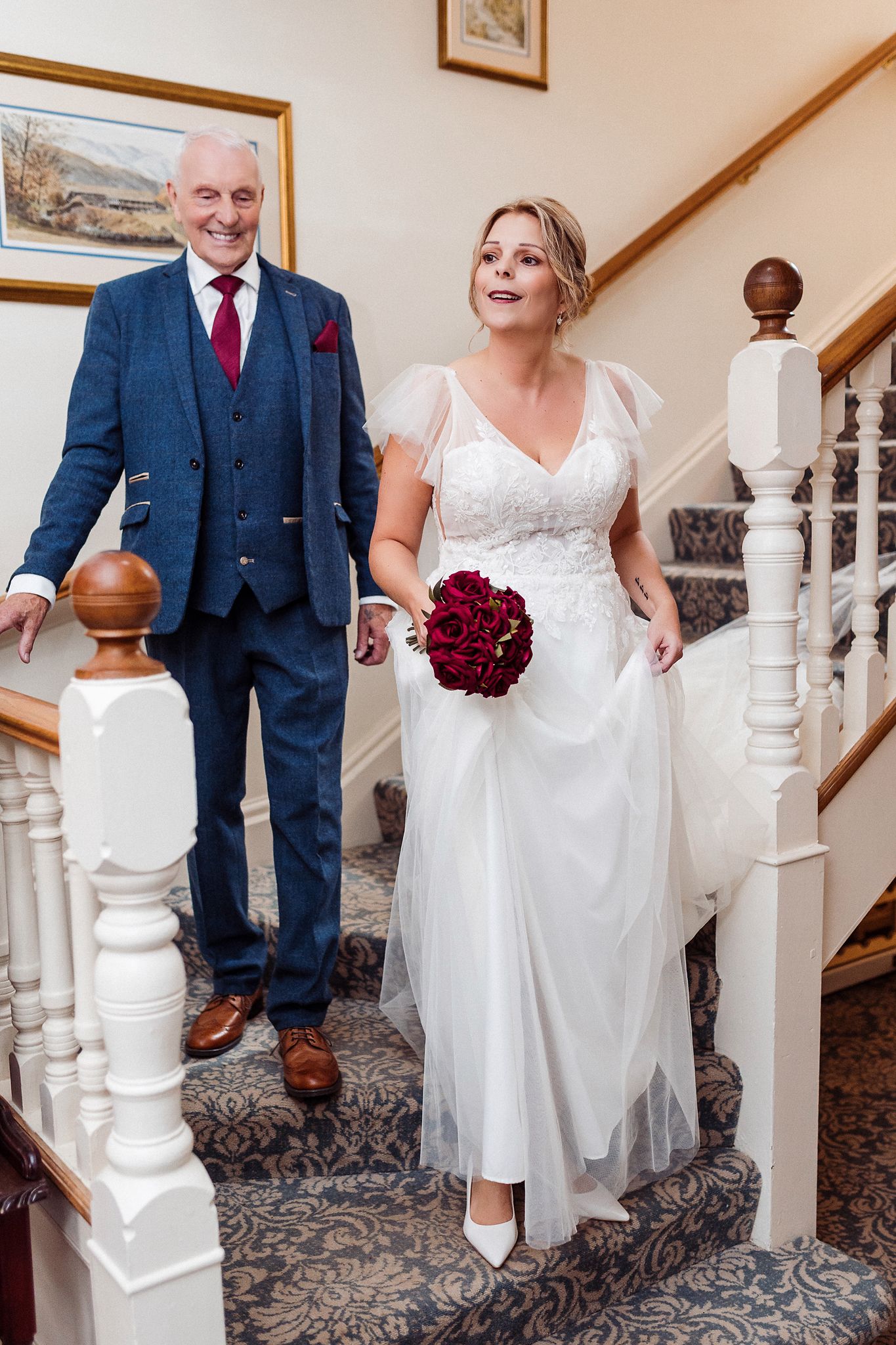 Wedding couple walks up carpeted staircase with white bannisters, bride holding deep red bouquet in elegant venue.