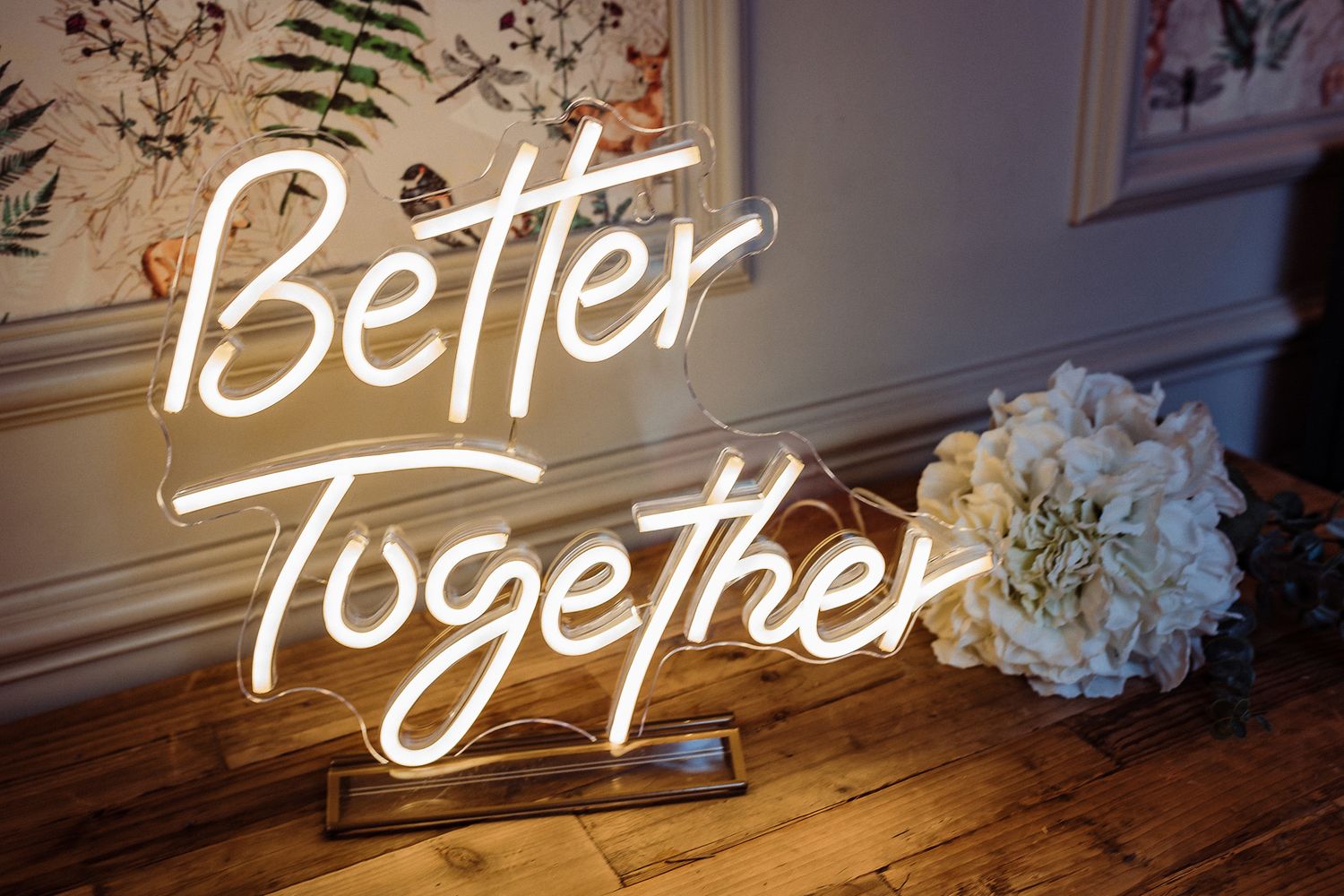 A series of illuminated neon signs spelling out 'Better Together' mounted on a white wall with floral decorations.