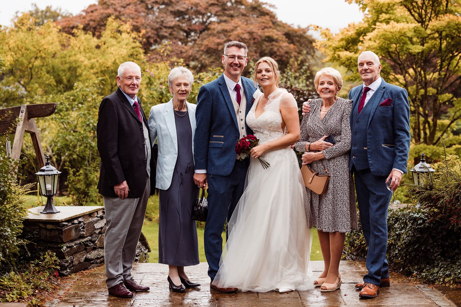Wedding party gathers for outdoor autumn photos in garden setting during golden hour sunset.